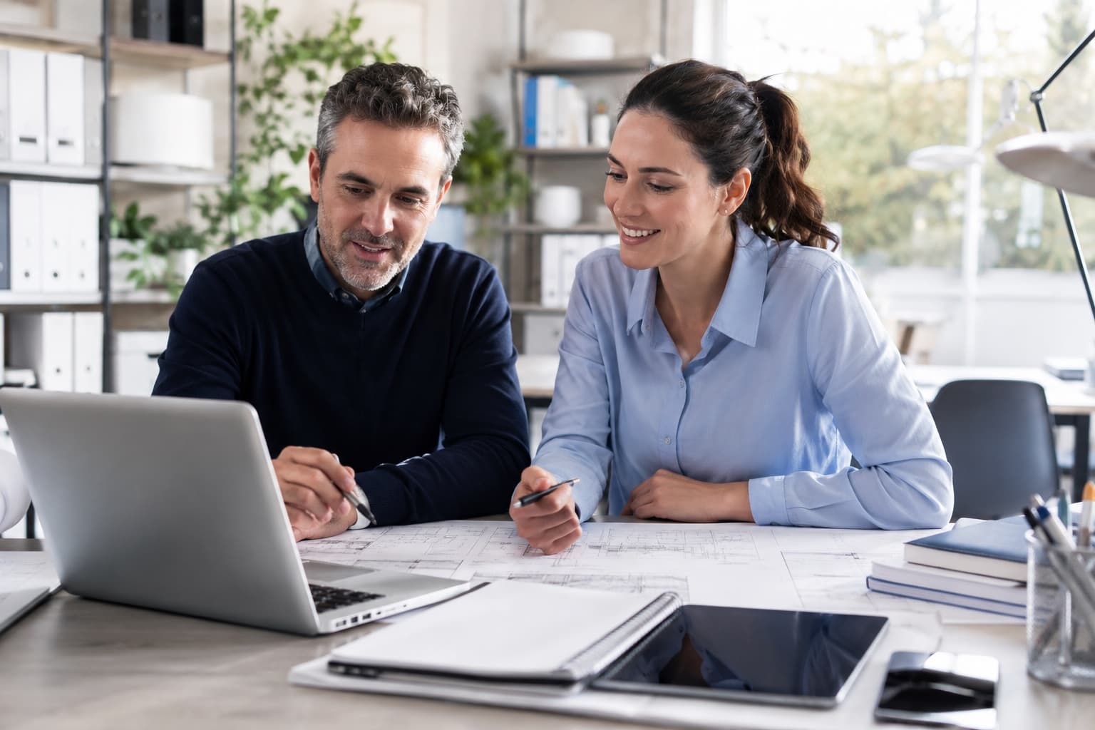Professionisti tecnici sorridenti lavorano su planimetrie e laptop per gestire clienti diretti